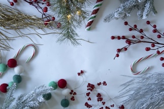 Festive decorations featuring pine branches, red berries, candy canes, and glittery gold and silver twigs are arranged on a white surface. Some elements are dusted with a white, snow-like texture, adding to the holiday feel. Various sizes of red, green, and white pom-poms are also visible.