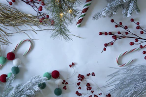 Festive decorations featuring pine branches, red berries, candy canes, and glittery gold and silver twigs are arranged on a white surface. Some elements are dusted with a white, snow-like texture, adding to the holiday feel. Various sizes of red, green, and white pom-poms are also visible.