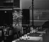 A reflection of a person walking is visible through a glass window, behind which a restaurant setting is displayed with neatly arranged tables and chairs. The scene also includes signage about boutique retail and office leasing opportunities, and an exit sign. The overall ambiance is a blend of urban and indoor environments.