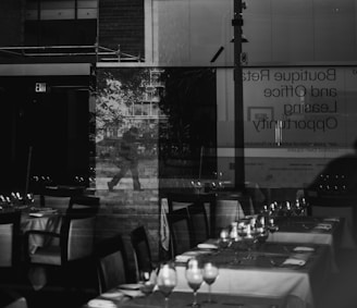A reflection of a person walking is visible through a glass window, behind which a restaurant setting is displayed with neatly arranged tables and chairs. The scene also includes signage about boutique retail and office leasing opportunities, and an exit sign. The overall ambiance is a blend of urban and indoor environments.