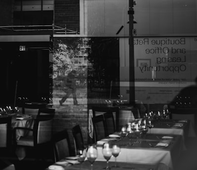 A reflection of a person walking is visible through a glass window, behind which a restaurant setting is displayed with neatly arranged tables and chairs. The scene also includes signage about boutique retail and office leasing opportunities, and an exit sign. The overall ambiance is a blend of urban and indoor environments.