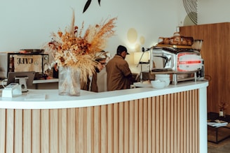 A modern and stylish cafe interior is focused on a white counter with wooden vertical paneling. A large glass vase filled with dried flowers is placed on the counter. Behind the counter, a barista is busy working with a large espresso machine. The decor is minimalistic with a warm, inviting atmosphere, enhanced by soft lighting and understated wall decorations.
