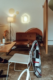 A cozy living room features a brown leather sofa against a white wall with a modern art design. A neon light shaped like a house is mounted above the sofa. To the left, a warm lamp illuminates a small table decorated with dried flowers. A wooden coffee table has a book and a mobile phone placed on it, and a checkered blanket is draped over a white metal chair.