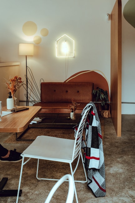 A cozy living room features a brown leather sofa against a white wall with a modern art design. A neon light shaped like a house is mounted above the sofa. To the left, a warm lamp illuminates a small table decorated with dried flowers. A wooden coffee table has a book and a mobile phone placed on it, and a checkered blanket is draped over a white metal chair.