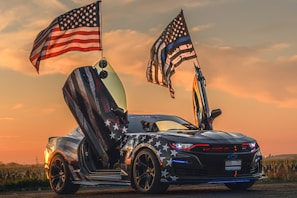 A sports car with American flag-themed decals is parked with its doors open, resembling a set of wings. Two large American flags are mounted on the back, waving in a breeze against a sunset background. The car has a sleek, modern design, emphasizing its powerful and aggressive aesthetic.
