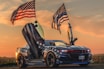A sports car with American flag-themed decals is parked with its doors open, resembling a set of wings. Two large American flags are mounted on the back, waving in a breeze against a sunset background. The car has a sleek, modern design, emphasizing its powerful and aggressive aesthetic.