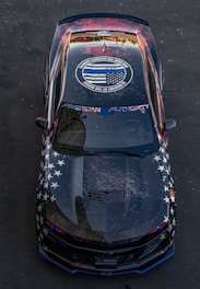 A black car with a customized paint job featuring white stars on the sides and a circular emblem on the roof. The emblem contains an American flag design with text surrounding it. The car's hood and roof display a weathered look with splatters of red paint, and the windshield has a decal with the words 'American Authority'.