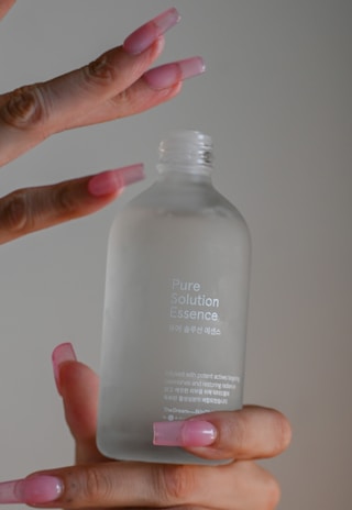 A close-up of a hand with long, pink acrylic nails holding a frosted glass bottle labeled 'Pure Solution Essence'. The bottle has a simple, elegant design with minimal branding in both English and Korean text.