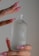 A close-up of a hand with long, pink acrylic nails holding a frosted glass bottle labeled 'Pure Solution Essence'. The bottle has a simple, elegant design with minimal branding in both English and Korean text.