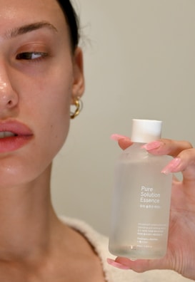 A person with a neutral expression is holding a clear bottle labeled 'Pure Solution Essence' with neatly manicured pink nails. The person has a clean, natural makeup look and an earring is visible.