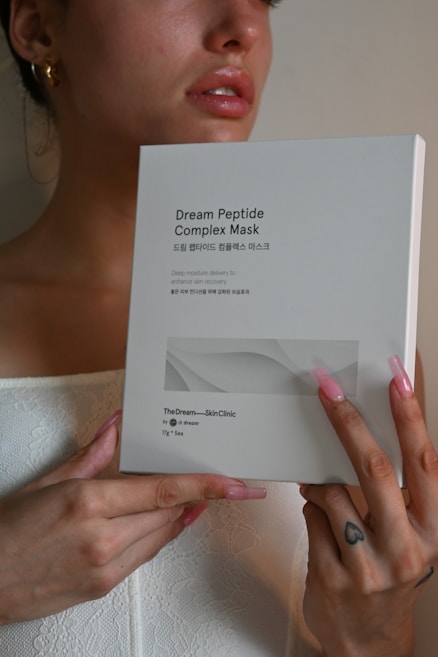 A person with long pink nails holds a box labeled 'Dream Peptide Complex Mask' against a neutral background. The box also has some Korean text and mentions 'TheDream – SkinClinic'. The person appears to be dressed in a white, textured fabric and has a tattoo on one finger.