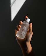 A hand holding a clear bottle labeled 'Pure Solution Essence,' with a minimalistic design. The background is dark with a geometric light pattern projected onto it.