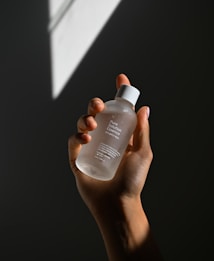 A hand holding a clear bottle labeled 'Pure Solution Essence,' with a minimalistic design. The background is dark with a geometric light pattern projected onto it.