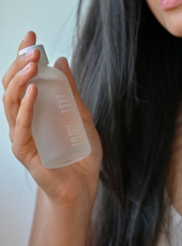 A hand with natural nails gently holds a frosted glass bottle labeled 'Pure Solution Essence' against a backdrop of long, dark hair. The focus is on the elegant bottle and the soft texture of the hair.