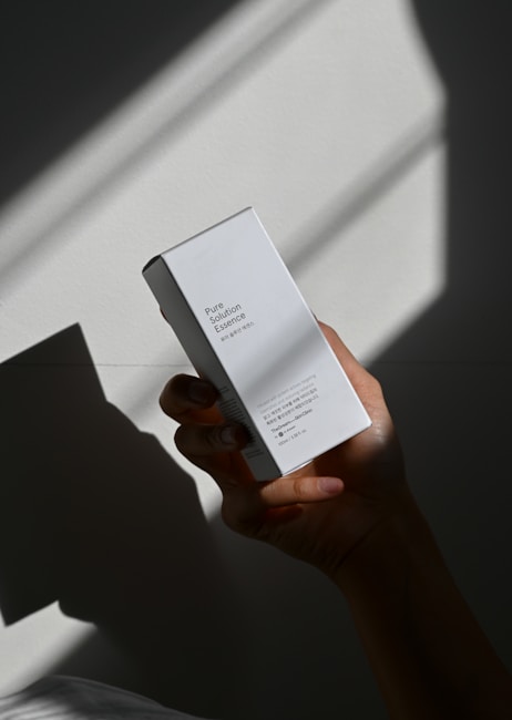 A hand holds a white box labeled 'Pure Solution Essence' against a backdrop with dramatic shadows. The box features minimal design with text in English and another language.