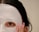 A close-up view of a person's face partially covered with a white sheet mask. One eye is visible through an opening in the mask, and a hand with long, manicured nails is gently touching the mask. The person's dark hair is partially visible.