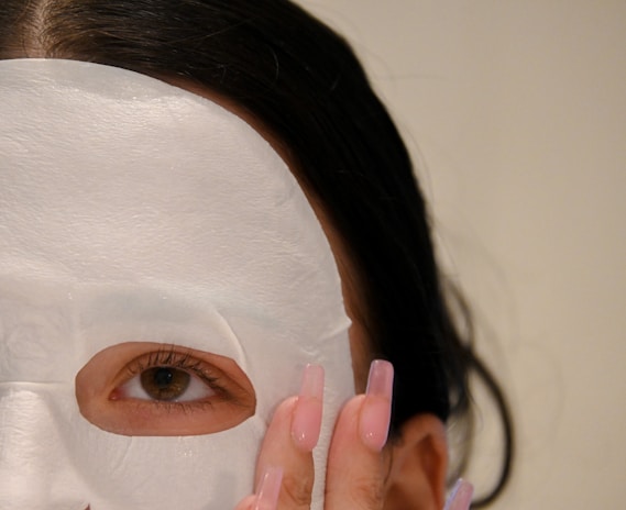 A close-up view of a person's face partially covered with a white sheet mask. One eye is visible through an opening in the mask, and a hand with long, manicured nails is gently touching the mask. The person's dark hair is partially visible.