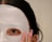 A close-up view of a person's face partially covered with a white sheet mask. One eye is visible through an opening in the mask, and a hand with long, manicured nails is gently touching the mask. The person's dark hair is partially visible.