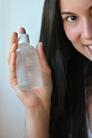A person with long dark hair smiles while holding a frosted glass bottle labeled 'Pure Solution Essence'. The hand and face are well-lit in natural light.
