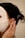 A person is wearing a white facial sheet mask with hands gently touching the side of their face. Their hair is styled back, adorned with pearl accessories. A small, gold earring is visible.