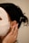 A person is wearing a white facial sheet mask with hands gently touching the side of their face. Their hair is styled back, adorned with pearl accessories. A small, gold earring is visible.