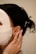 A person is wearing a white facial sheet mask with hands gently touching the side of their face. Their hair is styled back, adorned with pearl accessories. A small, gold earring is visible.