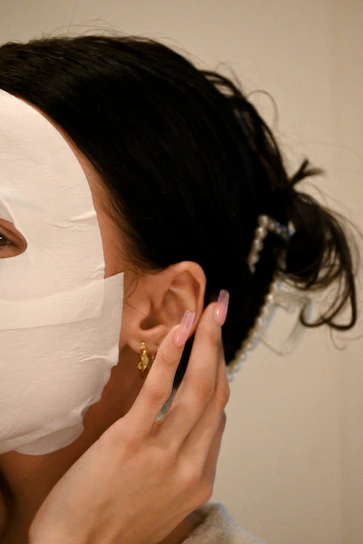 A close-up of a woman gently applying a hydrating Korean face mask, her skin glowing softly in natural light.