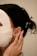 A person is wearing a white facial sheet mask with hands gently touching the side of their face. Their hair is styled back, adorned with pearl accessories. A small, gold earring is visible.