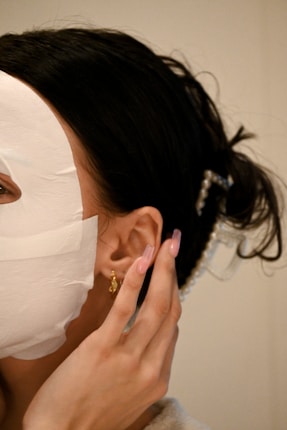 A person is wearing a white facial sheet mask with hands gently touching the side of their face. Their hair is styled back, adorned with pearl accessories. A small, gold earring is visible.