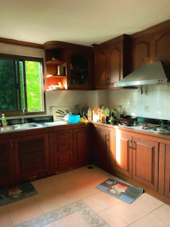 A warm, sunlit kitchen with wooden cabinets and countertops. A window on the left side lets in natural light, illuminating the area. Various cooking utensils and ingredients are arranged on the counter, and there are decorative mats on the tiled floor.