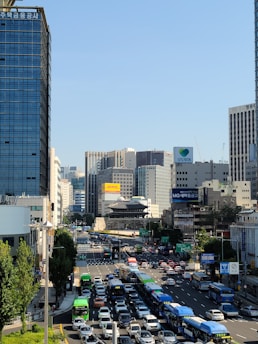 A busy urban street in a city surrounded by tall commercial buildings under a clear blue sky. The street is filled with traffic, including buses, cars, and trucks. On the horizon, a traditional Korean gate structure contrasts with the modern architecture.