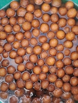 A collection of brown, round, ball-shaped desserts soaking in a syrupy liquid. The sweets appear glossy and are closely packed within a container.