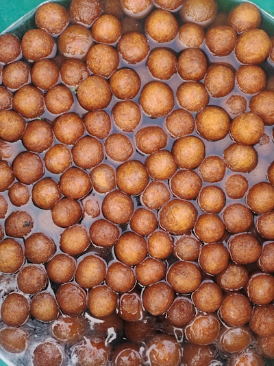 A collection of brown, round, ball-shaped desserts soaking in a syrupy liquid. The sweets appear glossy and are closely packed within a container.