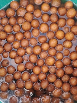 A collection of brown, round, ball-shaped desserts soaking in a syrupy liquid. The sweets appear glossy and are closely packed within a container.