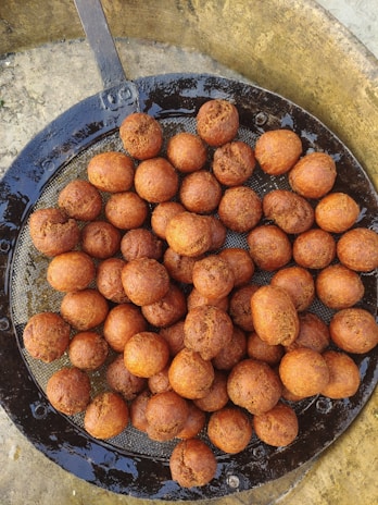 A collection of round, golden-brown fried dough balls arranged on a wire mesh tray set inside a round, metal container. The texture appears crispy and they are likely freshly cooked, glistening with a bit of oil.