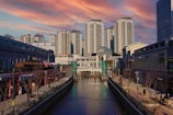 A modern urban area is depicted with several high-rise apartment buildings under a vibrant, colorful sky. The foreground features a canal bordered by walkways lined with shops and restaurants. A footbridge spans the canal further down.