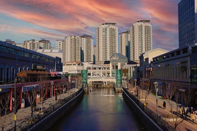 A modern urban area is depicted with several high-rise apartment buildings under a vibrant, colorful sky. The foreground features a canal bordered by walkways lined with shops and restaurants. A footbridge spans the canal further down.