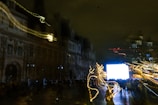 A night scene with a cityscape featuring a large historic building and a bright, illuminated screen. Blurred light trails of various colors, likely from moving lights or cameras, create a dynamic effect in the foreground, while silhouettes of people are visible in front of the building.
