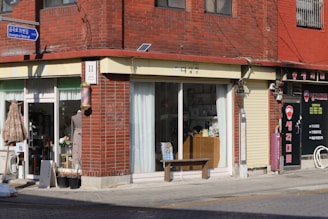 A street corner features a brick building with two storefronts. The left storefront has large glass windows with white curtains, displaying various items inside, and a sign with Korean text. A table with flowers, a mannequin, and plants are seen outside. The right storefront has a metal roll-up door, signage with red and white text, and additional decorations. The building is bathed in sunlight, casting shadows on the sidewalk.