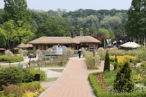 A tranquil garden scene with a brick pathway leading to a thatched-roof building surrounded by lush greenery and colorful flower beds. A fountain is centrally located. Tall trees and dense foliage line the background, and people can be seen walking around, enjoying the sunny day.