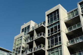 A modern apartment building with a concrete facade and large glass windows. The structure features multiple balconies with glass railings, and each level is stacked in a staggered formation. The sky is clear and blue, adding contrast to the architecture.