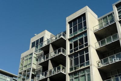 A modern apartment building with a concrete facade and large glass windows. The structure features multiple balconies with glass railings, and each level is stacked in a staggered formation. The sky is clear and blue, adding contrast to the architecture.