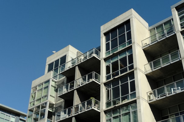 A modern apartment building with a concrete facade and large glass windows. The structure features multiple balconies with glass railings, and each level is stacked in a staggered formation. The sky is clear and blue, adding contrast to the architecture.