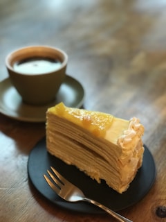 A slice of layered crepe cake topped with fruit and cream sits on a dark plate beside a cup of coffee on a wooden table. A small fork rests in front of the cake, and the soft lighting creates a warm and inviting atmosphere.
