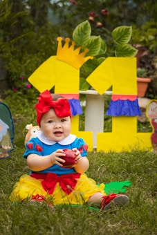 A baby dressed in a costume resembling a fairytale character is sitting on the grass, holding a red apple. The outfit features a blue top with red and yellow accents, including a large red bow on the head. In the background, there are colorful decorations with numbers and leaves, creating a festive atmosphere.