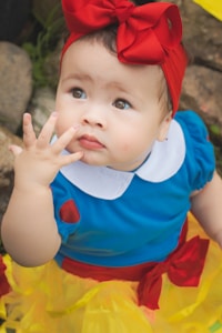 A young child wearing a vibrant blue top with a white collar, accessorized with a large red bow on their head. The child is looking upwards with a curious expression, surrounded by natural elements like rocks and greenery. The outfit is completed with a yellow skirt featuring red accents.
