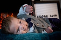 A baby is lying on a soft teal surface, dressed in a gray herringbone vest with wooden buttons, a white shirt, and a blue bow tie. The baby is wearing a red bracelet and appears to be in front of a sign with the name 'EDGAR LUIS'. In the background, there is a warm, glowing light resembling a fire.