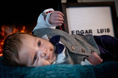 A baby is lying on a soft teal surface, dressed in a gray herringbone vest with wooden buttons, a white shirt, and a blue bow tie. The baby is wearing a red bracelet and appears to be in front of a sign with the name 'EDGAR LUIS'. In the background, there is a warm, glowing light resembling a fire.