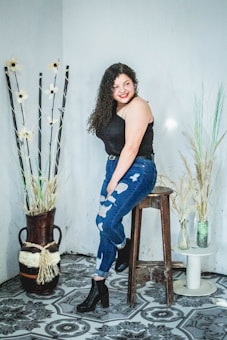 A woman with curly hair is sitting on a wooden stool, wearing a black sleeveless top and distressed jeans. She is smiling and looking off to the side. The floor features a decorative pattern, and there are decorative vases with dry plants and flowers on either side.
