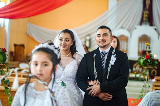 A couple is standing together in a church setting, dressed in wedding attire. The woman is wearing a white gown with lace details and a veil, accessorized with a pearl necklace. The man is in a black suit with a tie and decorative elements on his jacket. In front of them, a young girl is dressed in white, possibly a flower girl, with flowers in her hair. The background includes a decorated altar with drapes and floral arrangements.
